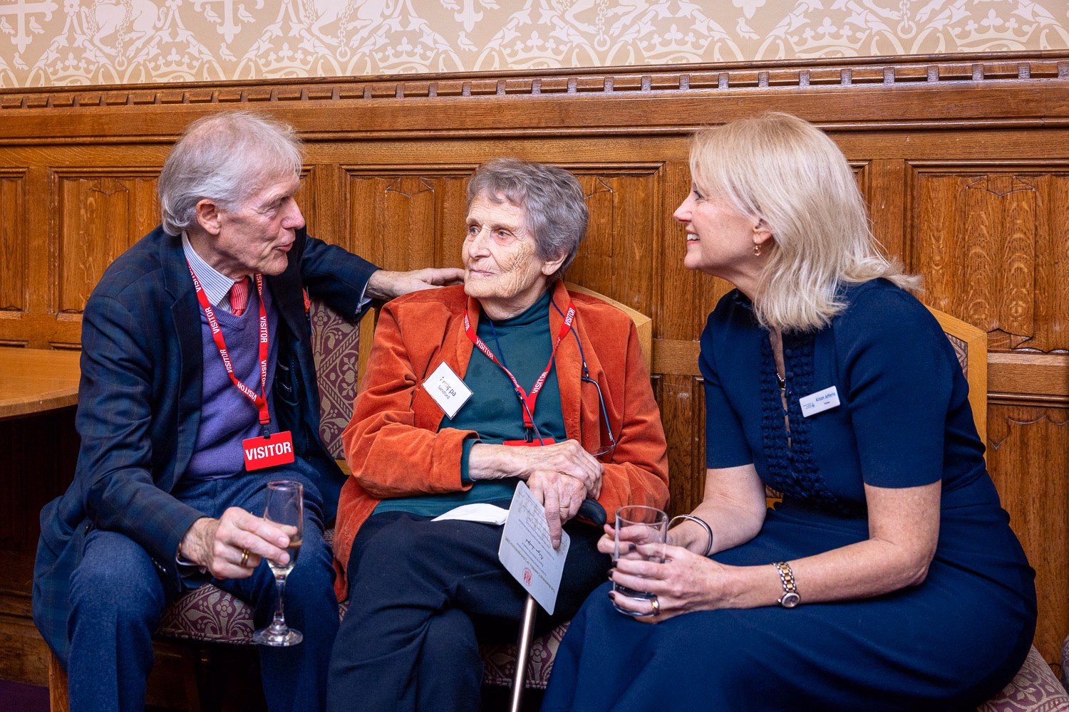 Man in suit chats to women in red jacket while a woman in a blue dress smiles